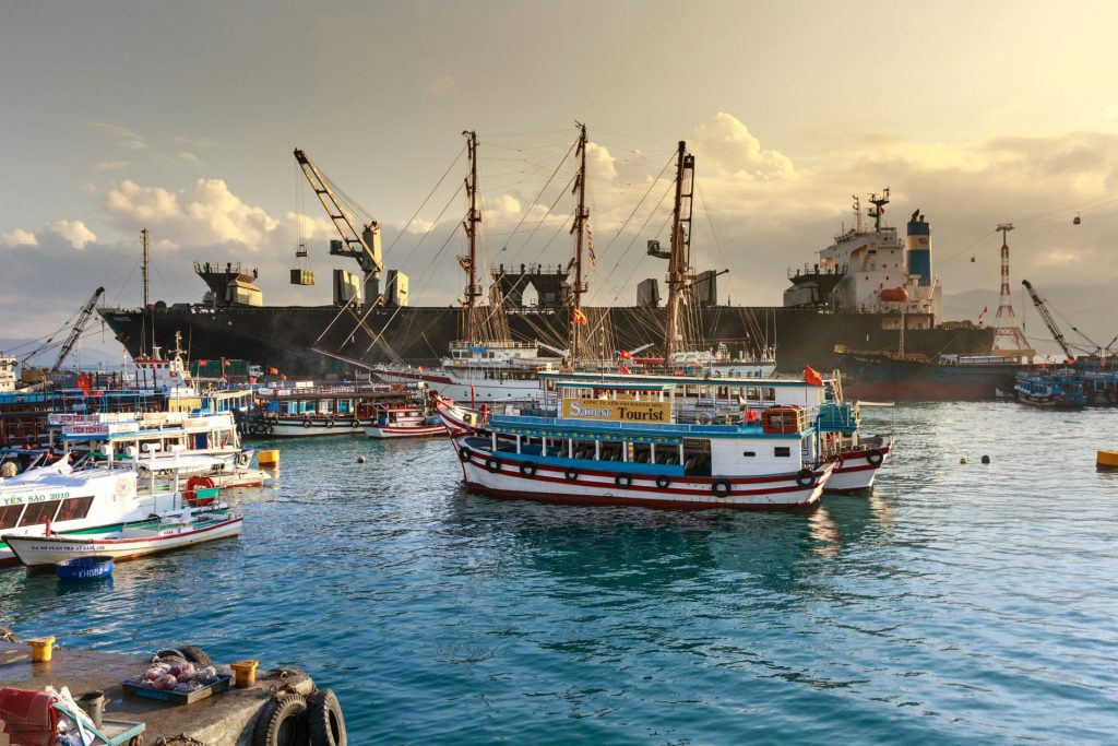 A busy harbor with cargo ships, cranes, and tourist boats under a cloudy sky.